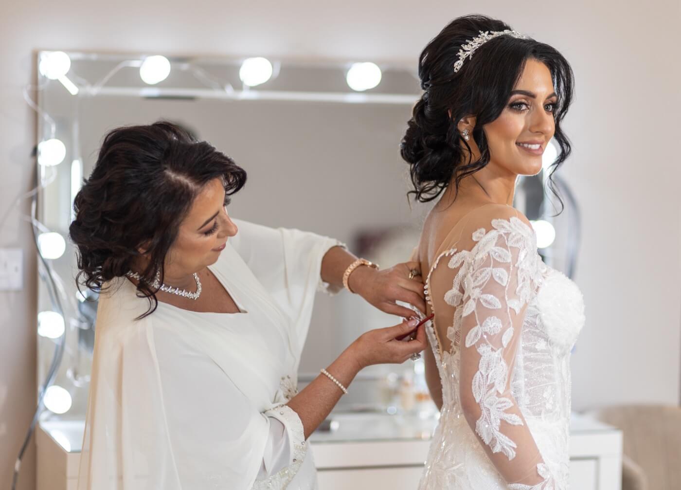 Bride smiling as her mother helps button her lace wedding dress in front of a lit mirror, Northern Ireland wedding photography by Annette Burke